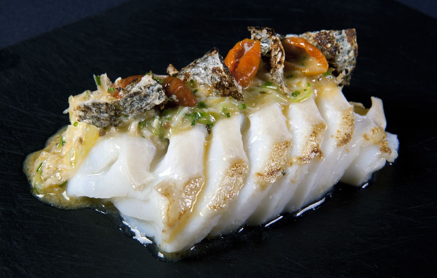 Bacalao presentado en lascas con salsa, tomates y trozos de piel dorada sobre un plato negro.